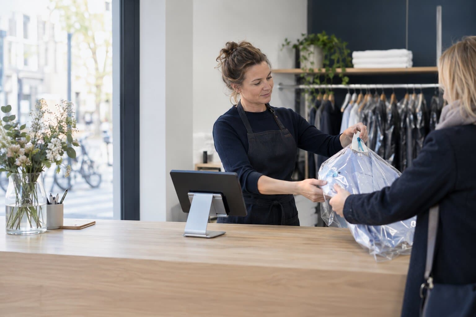 A dry cleaner hands a pressed garment to a customer across the counter, beside a tablet on a stand